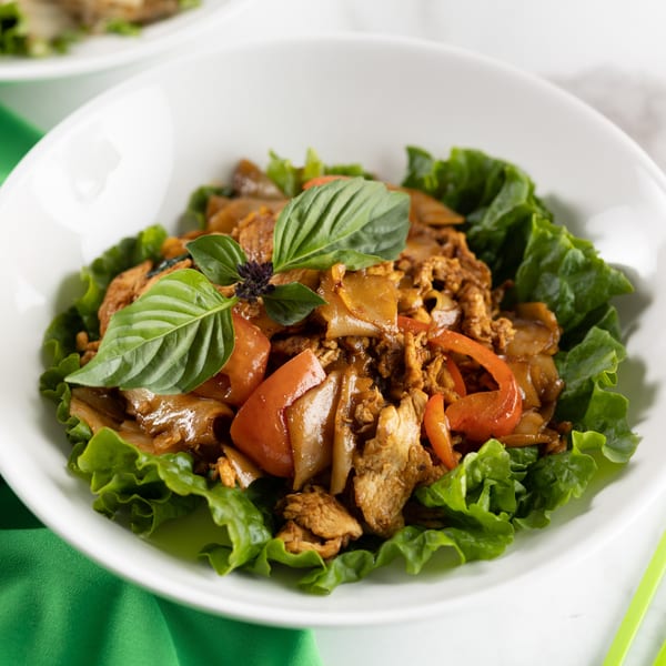 Thai drunken noodles with chicken, bell peppers, and basil, served in a lettuce-lined white bowl.
