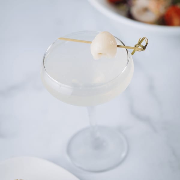 A clear martini glass filled with a light drink, garnished with a lychee on a bamboo skewer.