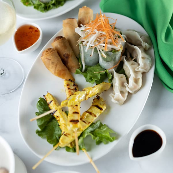 A white platter of Thai appetizers including spring rolls, fresh rolls, dumplings, and chicken satay skewers.