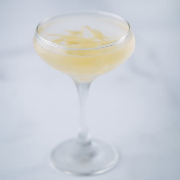 A pale yellow cocktail in a coupe glass, garnished with thin lemon zest, on a white marble surface.