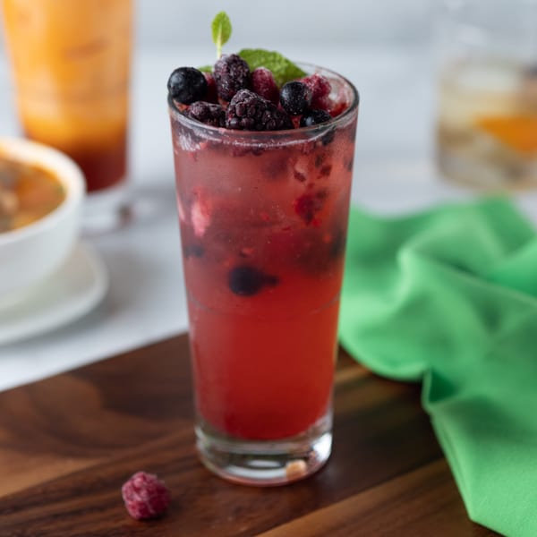 A vibrant red berry drink with mint on a wooden board, with other drinks blurred in the background.