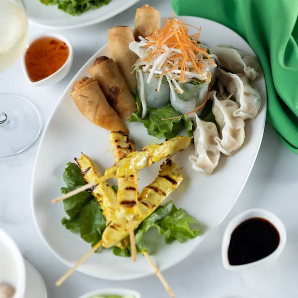 A white platter of Thai appetizers: spring rolls, fresh rolls, dumplings, and chicken satay skewers.