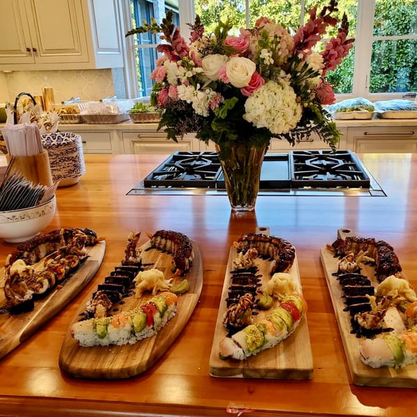 Sushi platters with various rolls and garnishes on a wooden kitchen island with flowers.