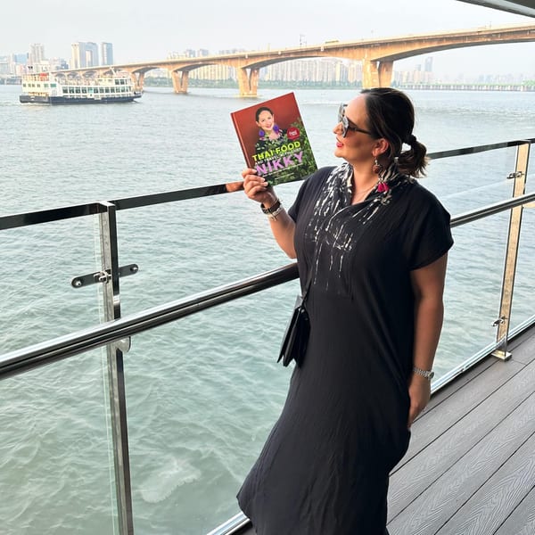 Chef Nikky holds her cookbook "Thai Food and Travel with Chef Nikky" on a boat overlooking a city.