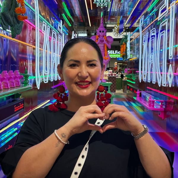 Woman in black shirt and red earrings making a heart shape with hands in a neon-lit Thai restaurant.