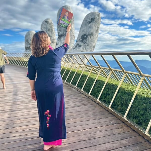 A woman in a blue dress holds up a "Thai Food and Travel with Nikki" book on a bridge.
