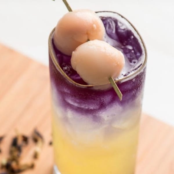A vibrant purple and yellow layered drink, garnished with two lychees on a skewer, on a wooden surface.