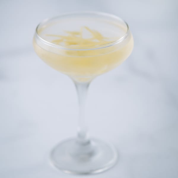 A coupe glass filled with a light yellow cocktail, garnished with thin lemon zest.