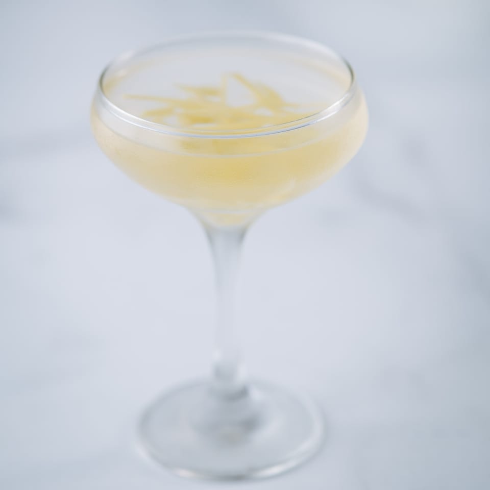 A coupe glass filled with a light yellow cocktail, garnished with thin lemon zest.