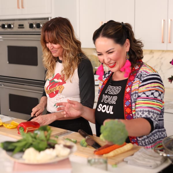 Two women smiling and chopping fresh vegetables in a bright kitchen, preparing a Thai meal.
