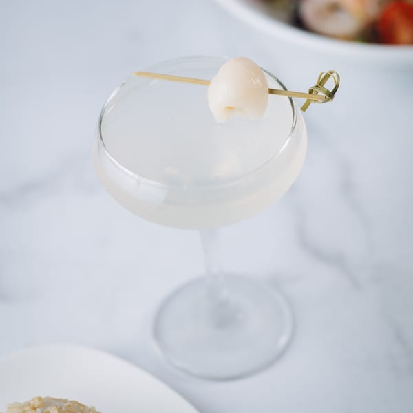 A lychee martini garnished with a single lychee on a skewer, on a white marble surface.