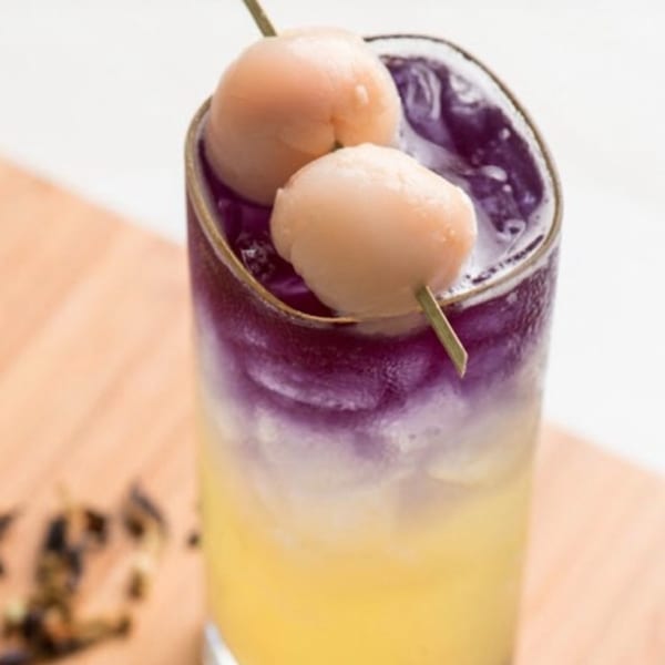 A tall glass of purple and yellow layered drink, garnished with two lychees on a skewer.