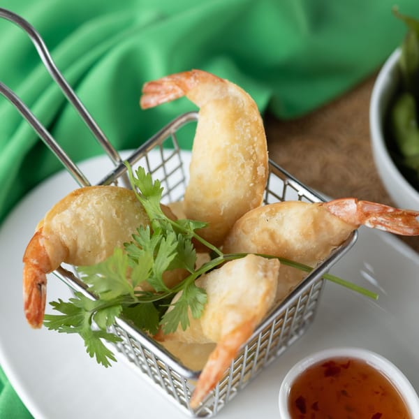 Crispy fried shrimp with visible tails, garnished with cilantro in a metal basket, with dipping sauce.