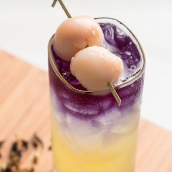 A vibrant purple and yellow layered drink, garnished with two lychees on a skewer.