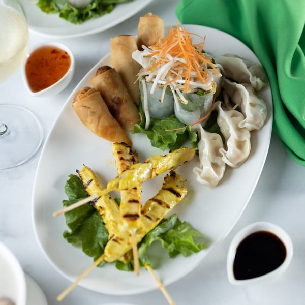 A white platter of Thai appetizers: chicken satay, spring rolls, fresh rolls, and dumplings with dipping sauces.