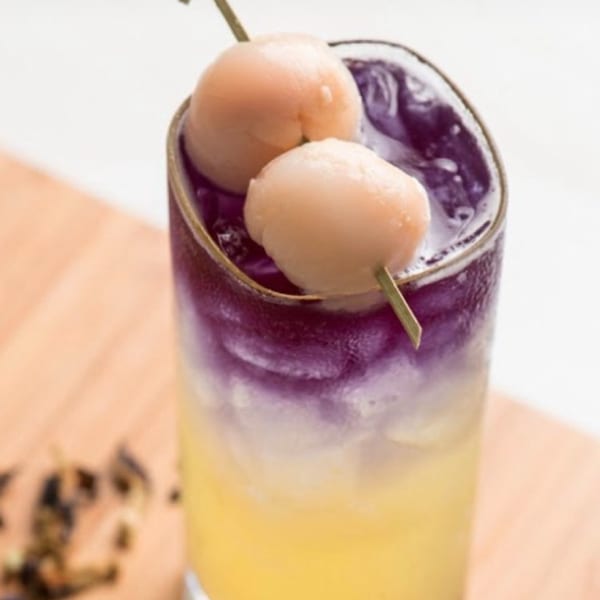 A vibrant yellow and purple layered drink, garnished with two lychees on a skewer.