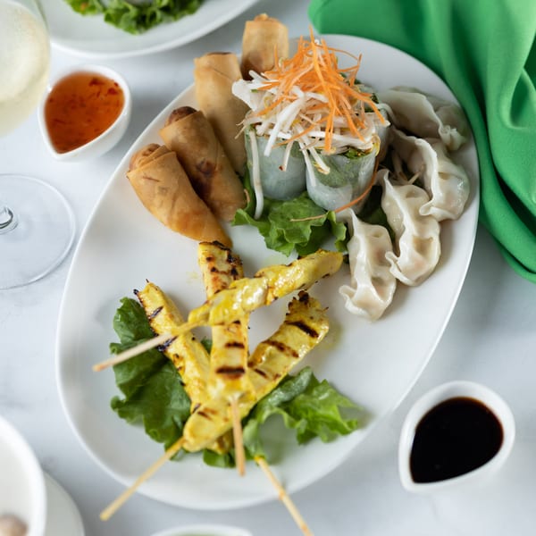A white platter of Thai appetizers: spring rolls, fresh rolls, chicken satay, and dumplings with dipping sauces.